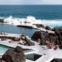 many people are sitting on the rocks near the water while others swim in the ocean