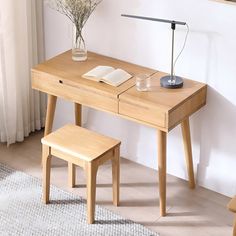 a wooden desk with a book, lamp and vase on it next to a window