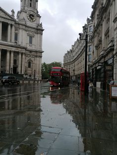 a red double decker bus driving down a rain soaked street in front of tall buildings