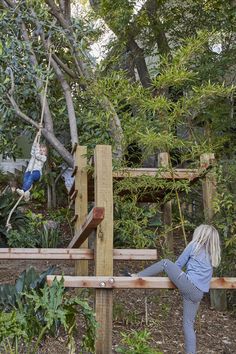 a woman standing on top of a wooden fence next to a lush green forest filled with trees