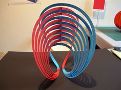 an abstract sculpture made out of red, blue and green strips on a table in front of a white wall