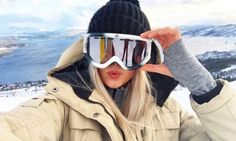 a woman wearing ski goggles while holding her hands up to her face with snow in the background