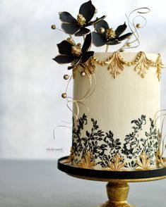 a white and gold decorated cake with black flowers on the top, sitting on a stand