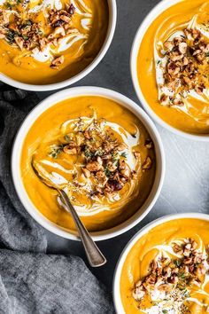 three bowls filled with carrot soup on top of a gray countertop next to spoons