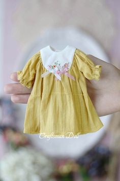 a hand holding a small doll wearing a yellow dress with pink flowers on it's chest