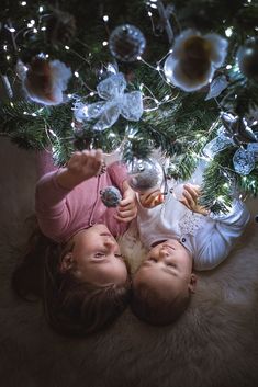 two young children laying under a christmas tree with ornaments hanging from it's branches