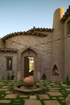 an outdoor courtyard with a fountain and lights strung from the ceiling over it, in front of a stone building