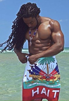 a man with long hair and no shirt on standing in front of the ocean looking down at his cell phone