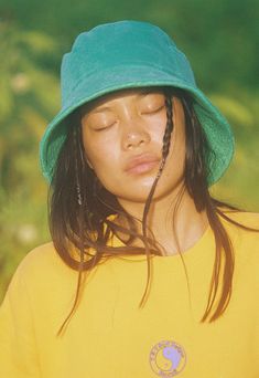 a woman wearing a yellow shirt and green hat with her eyes closed while holding a cell phone