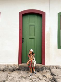 a woman sitting on the ground in front of a green door with red and white trim