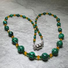 Two different sized and coloured Czech Crystal beads are combined with Green Malachite gemstone beads in this bright and colourful necklace. At each end of the necklace are five 4mm Malachite beads, then there are four 6mm Malachite's followed by three 8mm Malachite beads while finally in the centre are seven 10mm Malachite's. The Malachite beads are separated by an orange bicone Czech Crystal bead enclosed by two smaller green bicone Czech Crystal beads. All the metal work is Stainless Steel including the magnetic clasp and the earwires on the free matching earrings. Necklace and earring come enclosed within a gold foil fabric bag. Malachite ancient Greeks believe that Malachite reflects the heart of whoever hold it. Malachite is a protective stone that guards the bearer's heart from impu Beaded Malachite Round Bead Necklaces, Green Round Beads Spiritual Crystal Necklace, Healing Malachite Beaded Necklaces With Round Beads, Polished Malachite Round Beaded Necklaces, Green Crystal Necklaces With Polished Round Beads, Green Oval Beaded Jewelry 8mm, Green Crystal Necklaces With Round Gemstone Beads, Round Malachite Beaded Necklace, Green Crystal Necklace With Round Gemstone Beads