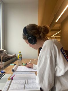 a woman sitting at a desk with headphones on and writing in a notebook next to a window