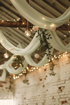 an indoor wedding venue with white drapes and greenery hanging from the ceiling, surrounded by string lights