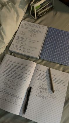 two notebooks and a pen sitting on top of a bed next to each other