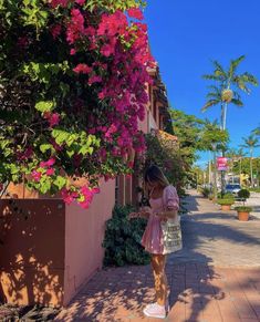 a woman standing on the sidewalk in front of a pink building with flowers growing out of it