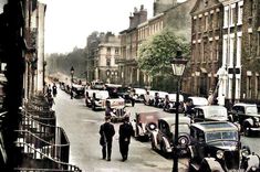 an old time street scene with cars and people walking on the sidewalk in front of buildings