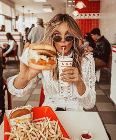 a woman sitting at a table with a sandwich and drink in her hand while holding a straw up to her mouth