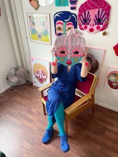 a woman sitting in a chair with a mask on her face and other art hanging above her head