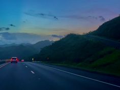 two cars driving down the road at dusk with mountains in the background and clouds overhead