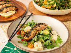 a white plate topped with fish next to a bowl filled with salad and chopsticks