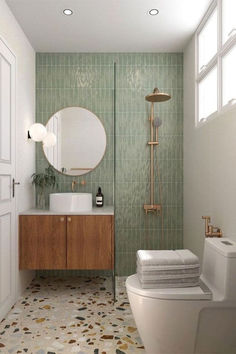 Deze moderne badkamer heeft een rustige uitstraling met groene tegels en een terrazzo-vloer. De ruimte bevat een zwevend houten meubel met een ronde opzetwastafel en een bijpassende ronde spiegel met gouden accenten. De douche heeft een elegante gouden regendouche. De verlichting komt van een stijlvolle wandlamp met twee glazen bollen. Het geheel is minimalistisch en warm, met een uitnodigende en georganiseerde sfeer. Bathroom Colour, Small Bathroom Interior, Restroom Decor, Downstairs Bathroom, Basement Bathroom