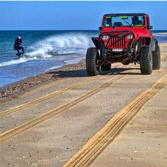 a red jeep driving down a sandy beach next to the ocean with a person in the background