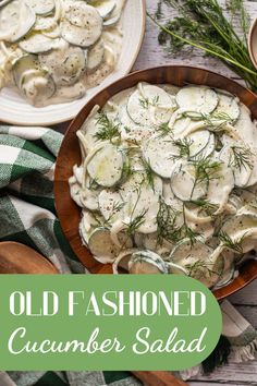 A wooden bowl filled with creamy cucumber salad sits on a white wooden table. It is sprinkled with fresh dill. There is more fresh dill nearby and a smaller white plate with a serving of the salad recipe sitting nearby.