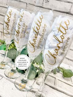 four champagne flutes with gold lettering and greenery in front of a white brick wall