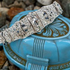 This elegant circa 1920s link bracelet is centered with a row of three (3), bead set, old European cut diamonds. The bracelet is also accented with one hundred sixty (160), bead set, old European/single cut diamonds, two (2), bezel set, cabochon cut synthetic sapphires and twenty (20), bezel set, calibre' cut synthetic sapphires. The gemstones are bordered with milgrain edging. The bracelet measures 18.4mm wide at the center and tapers to 5.0mm wide at the clasp. The bracelet measures 8-1/4 inches in length and finished with a hidden box clasp and safety chain. Art Deco Jewelry For Vintage Events, Heirloom Jeweled Wedding Bracelets, Heirloom Jeweled Bracelets For Wedding, Classic Formal Jewelry With Jewels, Luxury Collectible Cubic Zirconia Jewelry, Heirloom Style Jubilee Bracelet, Classic Diamond Accented Bracelet Jewelry, Vintage Diamond Bracelet With Hand-set Diamonds, Art Deco White Gold Jewelry With 17 Jewels