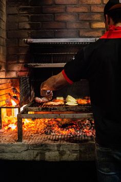 a man is cooking food on an outdoor grill in front of a brick wall and fire