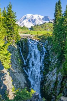 a waterfall in the middle of a forest with mountains in the background and trees around it