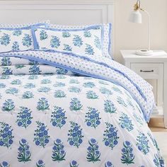 a white bed with blue flowers and polka dots on the comforter is next to a night stand