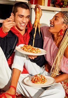 a man and woman sitting on the floor eating spaghetti from white plates with chopsticks in their hands