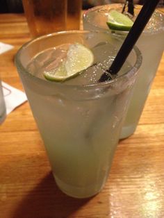 two glasses filled with ice and limes on top of a wooden table