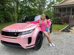 a woman leaning on the hood of a pink car in front of a house and trees