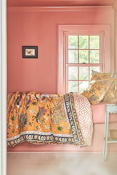 a bed sitting under a window next to a white chair and pink walls in a bedroom