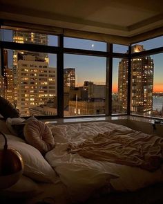 an unmade bed in front of a large window overlooking the city at night time