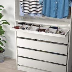 a white dresser with drawers and clothes hanging on the wall next to a potted plant