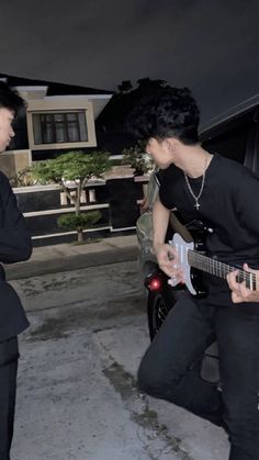 two men are playing guitars in front of a car and one is wearing a black suit