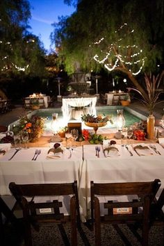 an outdoor dinner table set for two with candles and lights on the water fountain in the background