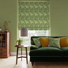 a living room with green walls and yellow pillows on the couch, next to a window covered in roman shades