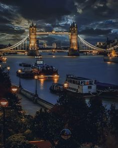 the tower bridge is lit up at night with lights on and boats in the water below