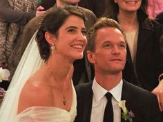 a bride and groom smile as they sit in front of their guests at a wedding