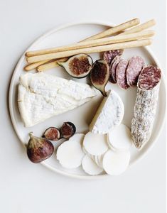 a white plate topped with different types of cheese and fruit next to breadsticks