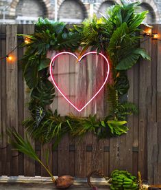 a heart - shaped neon sign hangs on a wooden fence surrounded by tropical plants and bananas