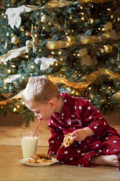 a little boy sitting on the floor drinking milk from a glass while eating cookies with a straw in front of a christmas tree