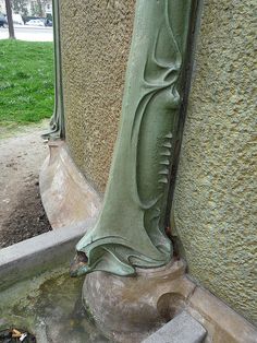 a water fountain in the corner of a building next to a sidewalk and grassy area