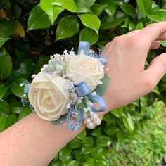 a wrist corsage with white roses and blue ribbon is shown in front of green leaves