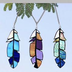 three stained glass feathers hanging from a chain on a tree branch in front of a blue sky