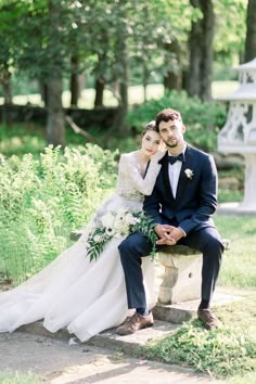 a bride and groom are sitting on a stone bench in the park, posing for a photo